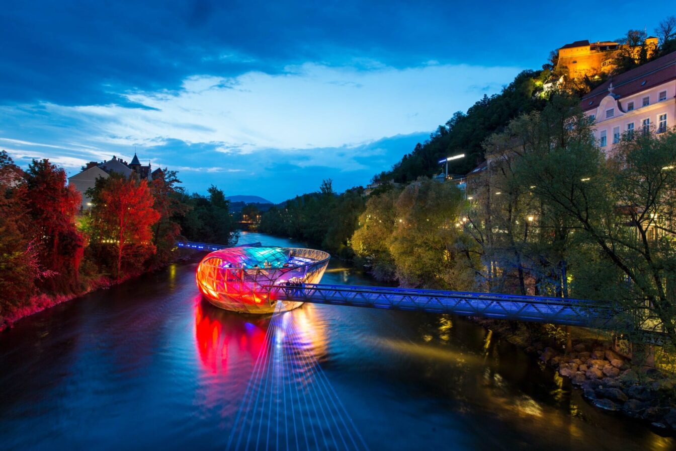 Ansicht der Murinsel bei Dämmerung. Der Fluss ist bereits dunkelblau, die Insel erstrahlt in vor allem rotem Licht, aber darunter mischen sich Gelb und Blau.