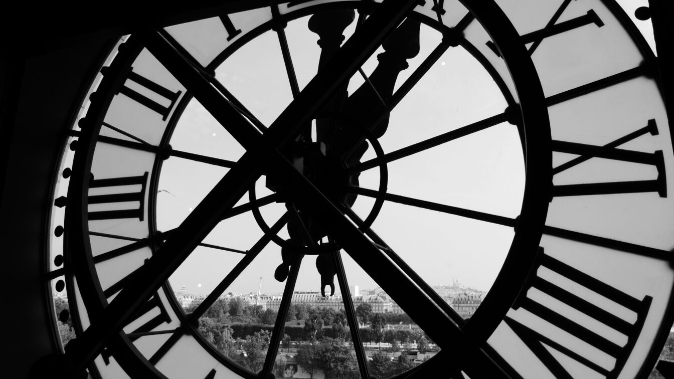 (c) Murray Campbell | Unsplash Blick durch eine große, historische Uhr mit römischen Ziffern, die von innen fotografiert wurde. Im Hintergrund ist eine verschwommene Stadtlandschaft zu sehen, möglicherweise durch das Glas verzerrt. Das Schwarz-Weiß-Foto betont die Details der Uhr und die Silhouette der dahinterliegenden Gebäude.