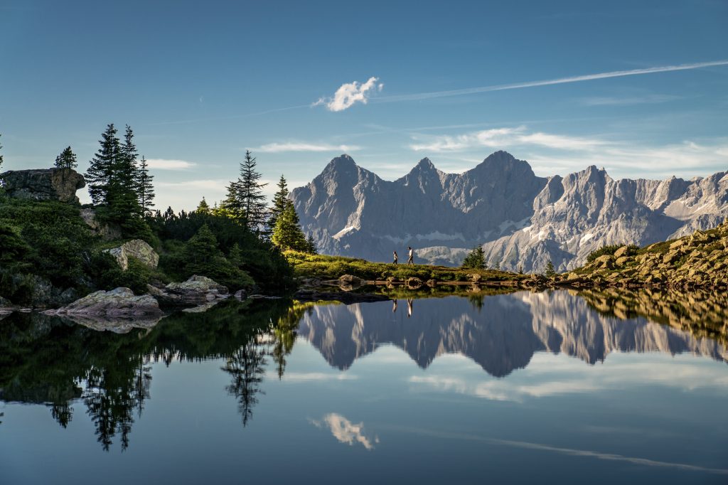 Glasklarer Bergsee mit perfekter Spiegelung der markanten Dachstein-Bergkette im Wasser. Im Vordergrund umrahmen Tannen, Felsen und Almwiesen den ruhigen See. Die Szenerie liegt unter klarem Himmel in der Morgen- oder Abendsonne, die das felsige Panorama in warmes Licht taucht.