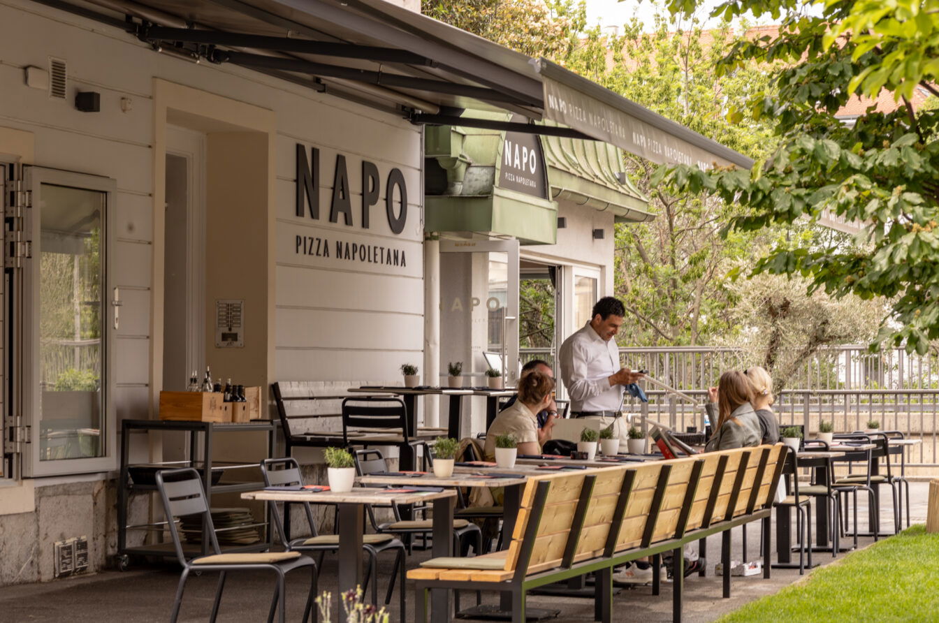 Außenbereich der Napo Pizzeria in Graz mit modernem, gemütlichem Ambiente. Gäste sitzen an Holzbänken und Metallstühlen an gedeckten Tischen, während ein Kellner die Bestellung aufnimmt. Das Restaurant liegt im Grünen und ist mit klarer Beschilderung „NAPO Pizza Napoletana“ gekennzeichnet. Pflanzen und dezente Deko schaffen eine einladende Atmosphäre.