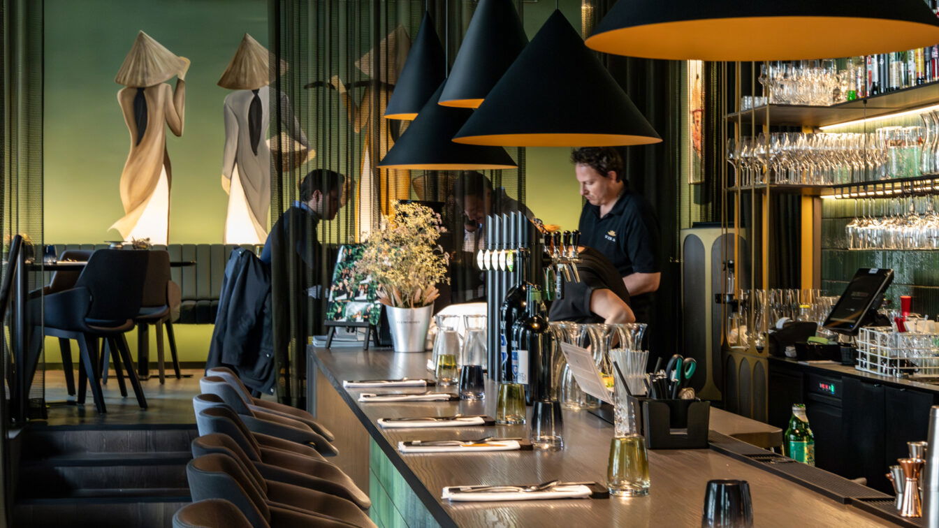 (c) Sonja Koller | 1000things Stilvoll eingerichteter Innenraum des Restaurants Vina in Graz. Im Vordergrund eine elegante Bar mit gedeckten Plätzen, Flaschen und Gläsern. Schwarze, konisch geformte Lampen hängen über der Theke. Im Hintergrund bedienen Mitarbeitende an der Bar, während Gäste an Tischen im erhöhten Sitzbereich sitzen. Die grüne Wand mit großem Wandgemälde zweier stilisierter Figuren in traditionellen vietnamesischen Kleidern verleiht dem Raum ein besonderes Ambiente.
