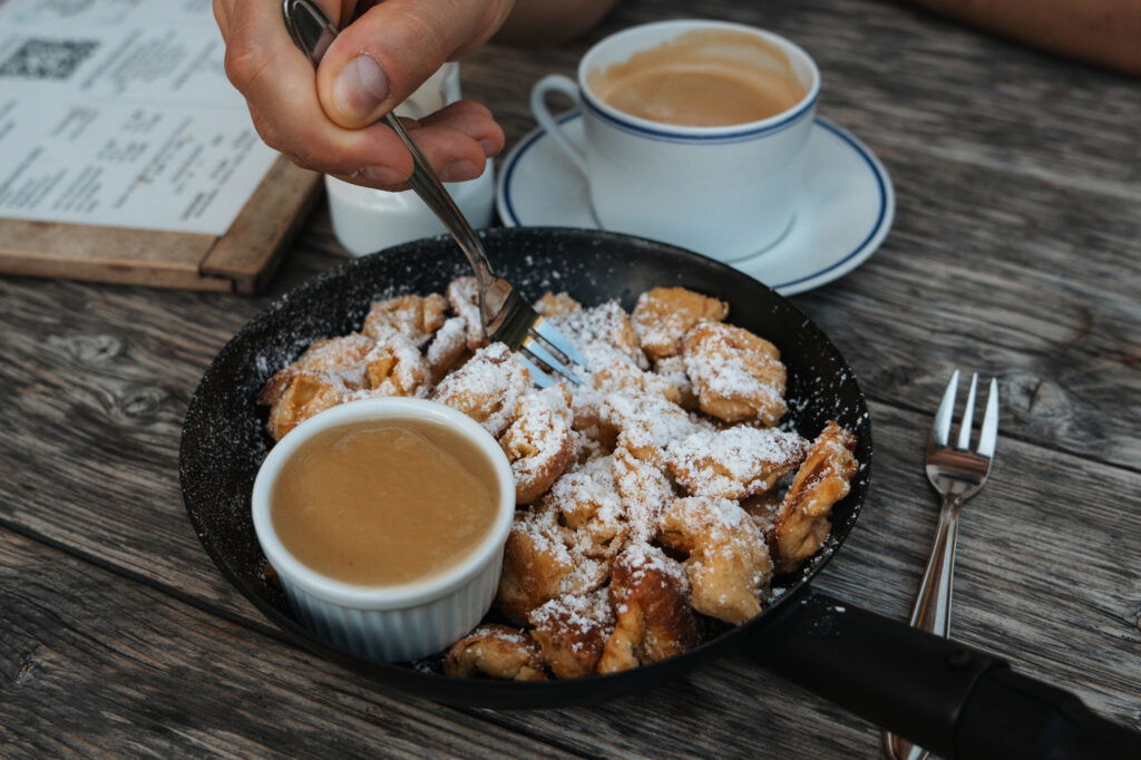 Im Bild wird eine Hand gezeigt, die mit einer Gabel Kaiserschmarrn aus einer Pfanne isst, der mit Puderzucker bestäubt ist. Daneben steht ein kleines Schälchen mit Apfelmus sowie eine Tasse Kaffee auf einem Holztisch.