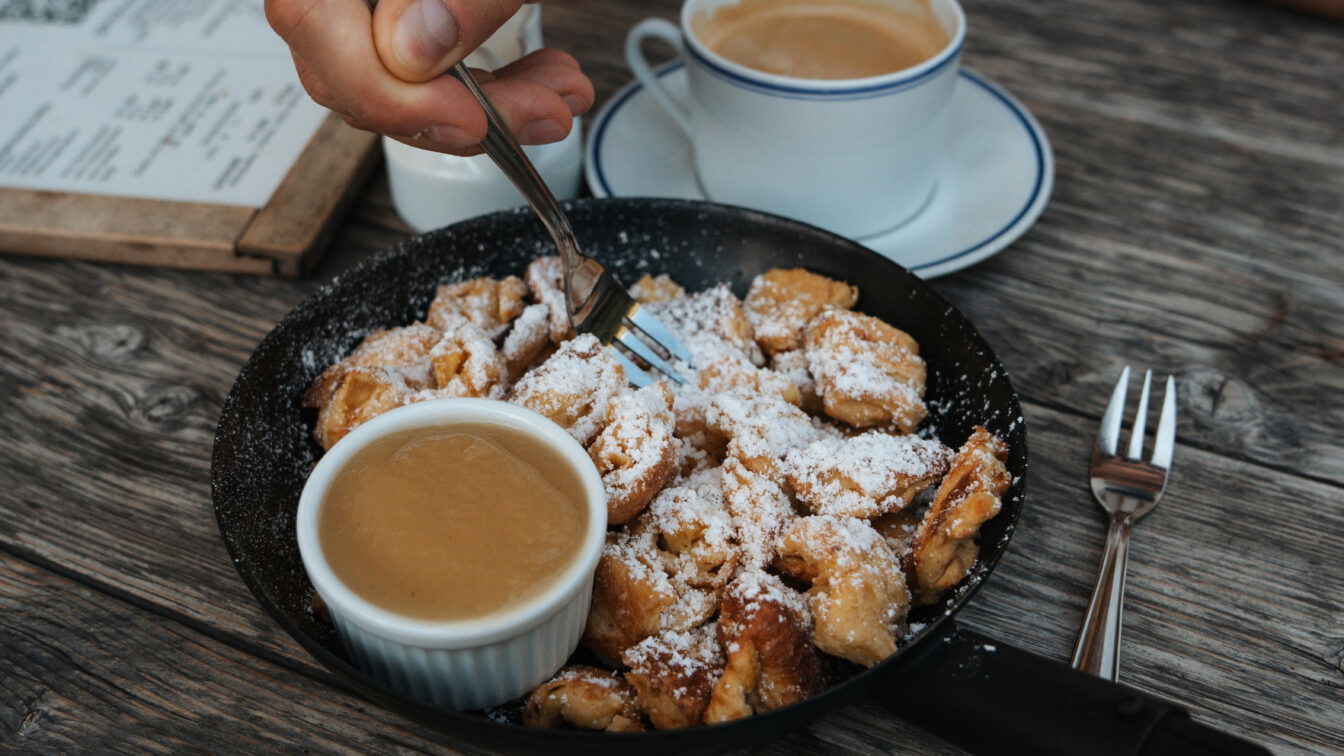 (c) Michelle Doninger Im Bild wird eine Hand gezeigt, die mit einer Gabel Kaiserschmarrn aus einer Pfanne isst, der mit Puderzucker bestäubt ist. Daneben steht ein kleines Schälchen mit Apfelmus sowie eine Tasse Kaffee auf einem Holztisch.