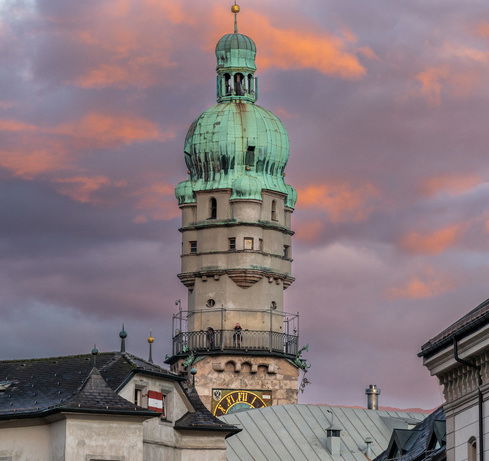 Innsbruck Stadtturm, Das Bild zeigt den Stadtturm von Innsbruck mit seiner markanten, grünen Kupferkuppel. Der Turm erhebt sich über die Dächer der umliegenden Gebäude und wird von einem dramatischen Himmel mit orangefarbenen und violetten Wolken bei Sonnenuntergang hinterlegt.