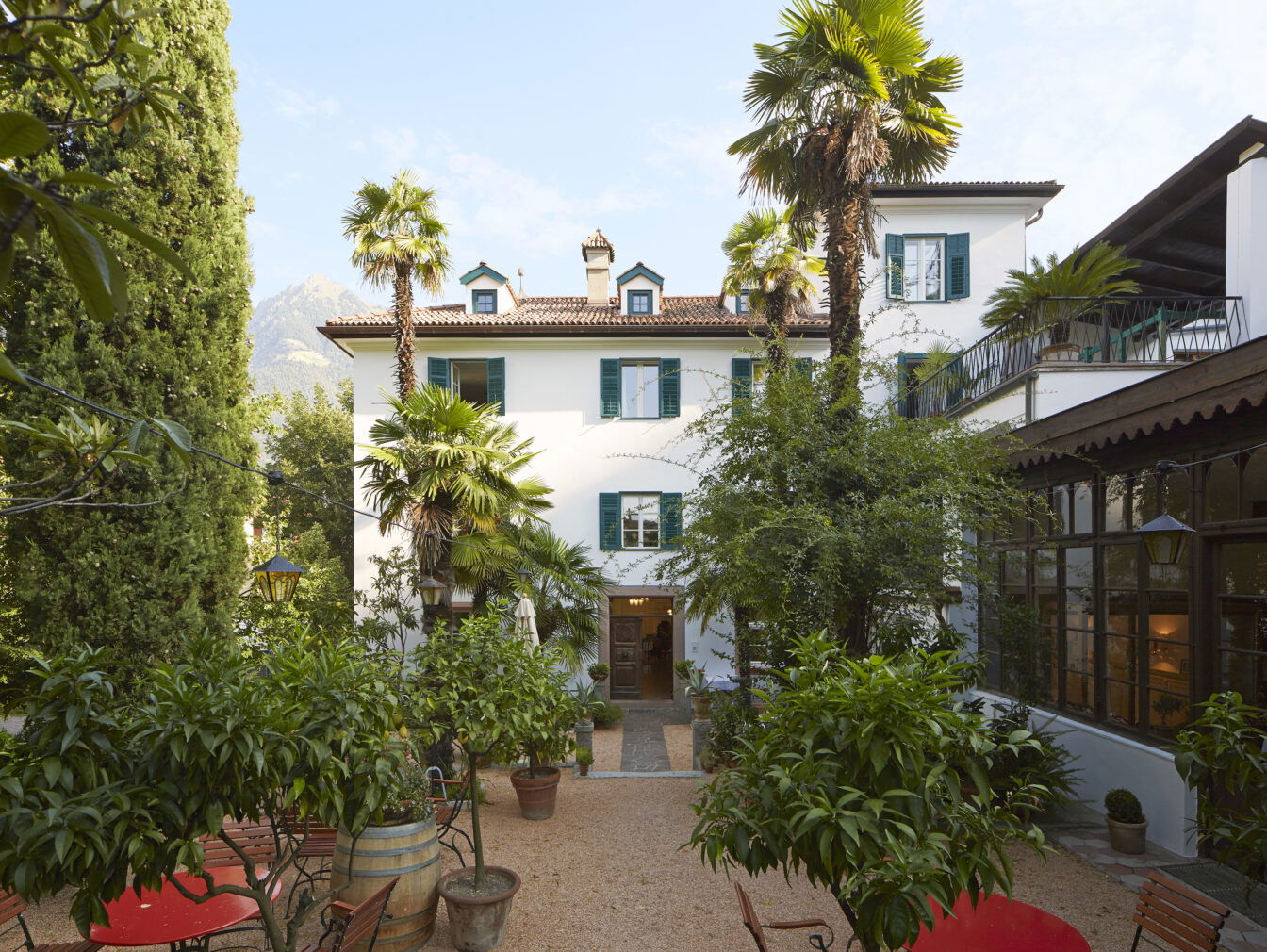 Das Ottmanngut, ein charmantes historisches Gebäude im mediterranen Stil, umgeben von Palmen und üppiger Vegetation. Im Vordergrund stehen Gartenmöbel und Töpfe mit Orangenbäumen, die eine einladende, ruhige Atmosphäre schaffen. Im Hintergrund sind Berge und blauer Himmel zu sehen.