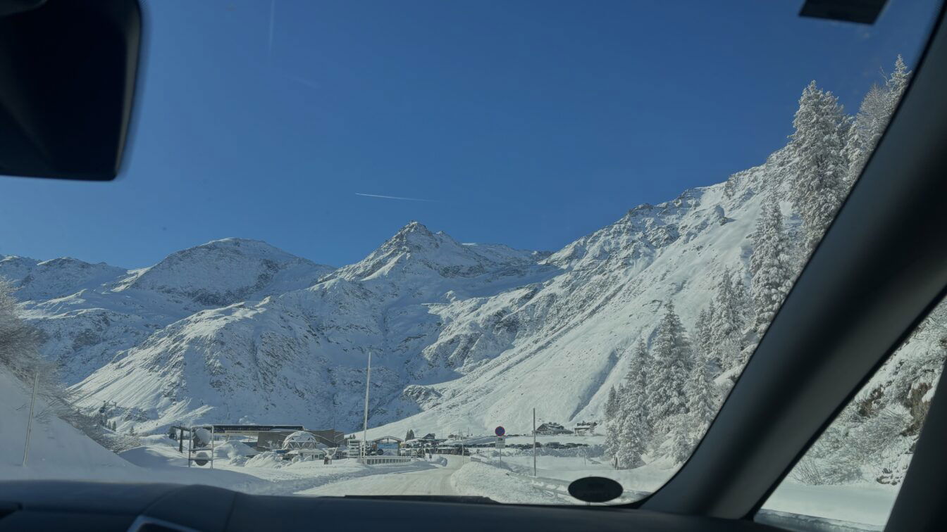 (c) Sonja Koller | 1000things Das Bild zeigt eine beeindruckende Berglandschaft, aufgenommen aus einem Auto heraus. Im Vordergrund ist die Windschutzscheibe des Fahrzeugs sichtbar, während sich die schneebedeckten Berge und Bäume im Hintergrund erstrecken. Der Himmel ist klar und strahlend blau, was einen schönen Kontrast zu der weißen Schneedecke bildet. Es scheint eine winterliche Bergstraße zu sein, mit einem Gebäude und einigen Fahrzeugen im Talbereich. Das Bild vermittelt eine ruhige und majestätische Atmosphäre eines Bergortes.
