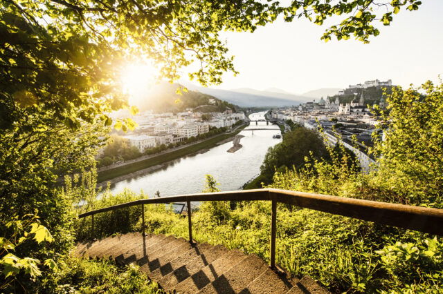 Sonnenuntergang über Salzburg mit Blick auf die Altstadt und die Festung Hohensalzburg. Eine von Bäumen umrahmte Treppe führt hinab zu einem malerischen Ausblick auf Salzburg. Die Sonne taucht die Stadt in goldenes Licht, während die Salzach durch das Stadtzentrum fließt. Brücken verbinden die beiden Ufer, und im Hintergrund thront die imposante Festung Hohensalzburg über den historischen Gebäuden. Ein idyllischer und stimmungsvoller Moment inmitten der Natur. Sonnenuntergang über Salzburg mit Blick auf die Altstadt und die Festung Hohensalzburg. Eine von Bäumen umrahmte Treppe führt hinab zu einem malerischen Ausblick auf Salzburg. Die Sonne taucht die Stadt in goldenes Licht, während die Salzach durch das Stadtzentrum fließt. Brücken verbinden die beiden Ufer, und im Hintergrund thront die imposante Festung Hohensalzburg über den historischen Gebäuden. Ein idyllischer und stimmungsvoller Moment inmitten der Natur.