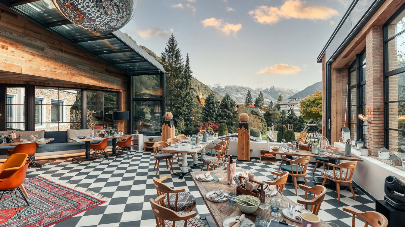 (c) Sendlhofers Sendlhofers in Salzburg. Moderner Wintergarten eines Restaurants mit großen Fensterfronten und Bergpanorama im Hintergrund. Der Raum wirkt gemütlich und einladend, mit Holzmöbeln, kariertem Boden und einer großen Diskokugel an der Decke. Der Blick reicht über grüne Nadelbäume und Berge unter blauem Himmel mit wenigen Wolken.