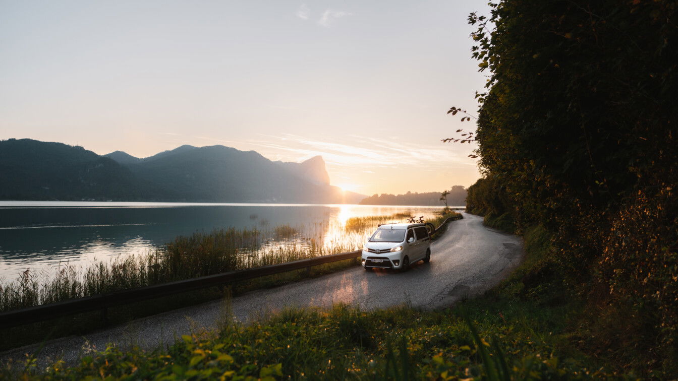 Foto Oberösterreich Tourismus GmbH/Moritz Ablinger: Fahrender Camping Van am Mondsee bei Sonnenuntergang.