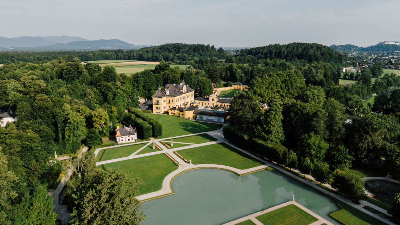 (c) SalzburgerLand Tourismus Panoramaaufnahme von Schloss Hellbrunn in Salzburg mit prächtiger Parkanlage, barockem Lustschloss und Wasserspielen, eingebettet in die grüne Naturlandschaft.