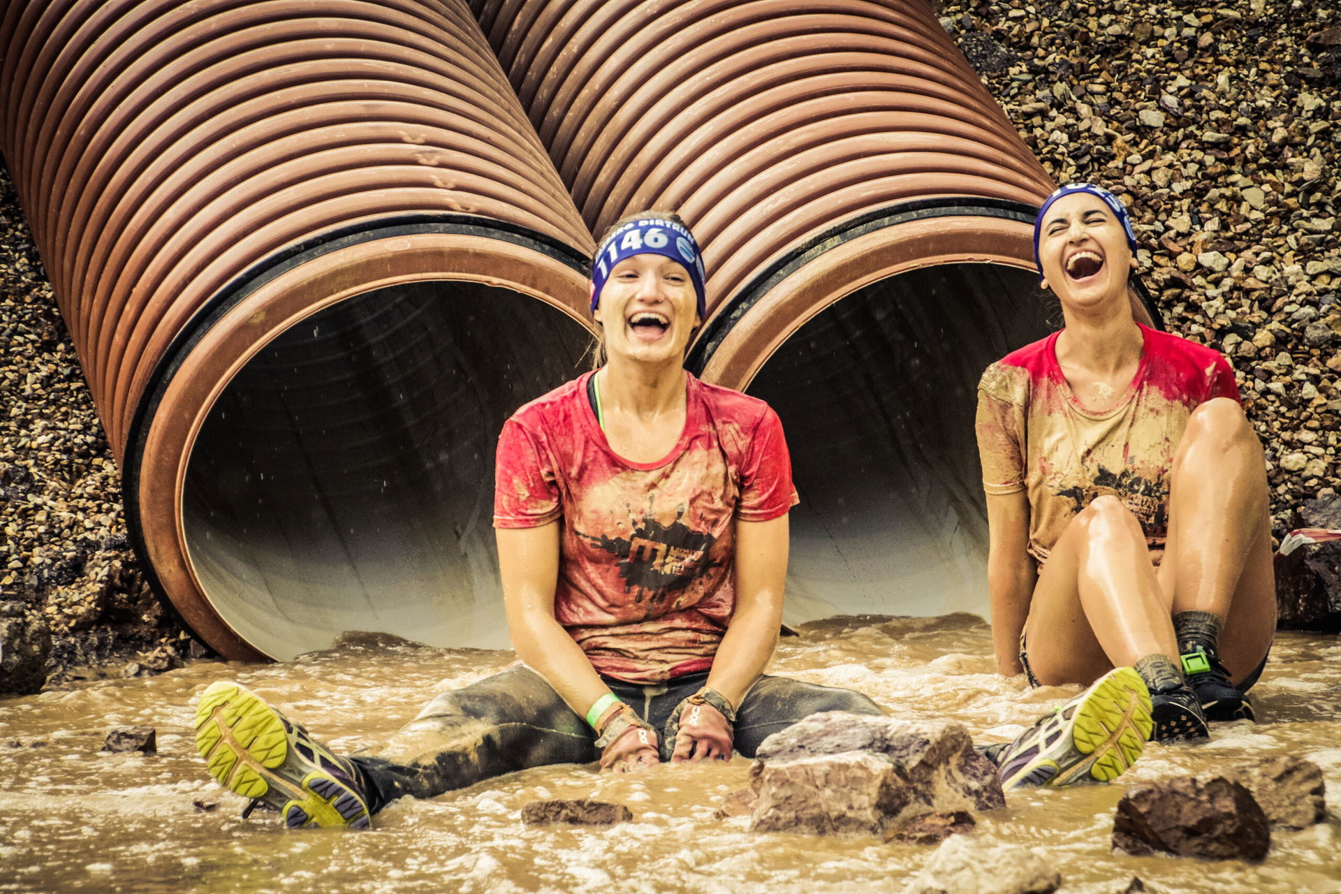 Zwei Frauen sitzen im Schlamm und lachen herzlich beim Krone Erzberg Adventure Day, hinter ihnen Metallrohre.