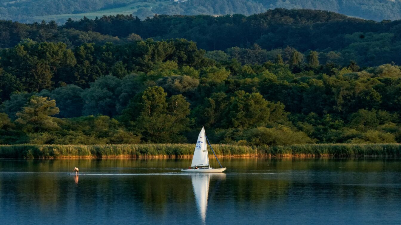 (c) John Aapps Unsplash Segelboot auf dem Chiemsee bei Sonnenuntergang vor bewaldeter Hügellandschaft in Bayern