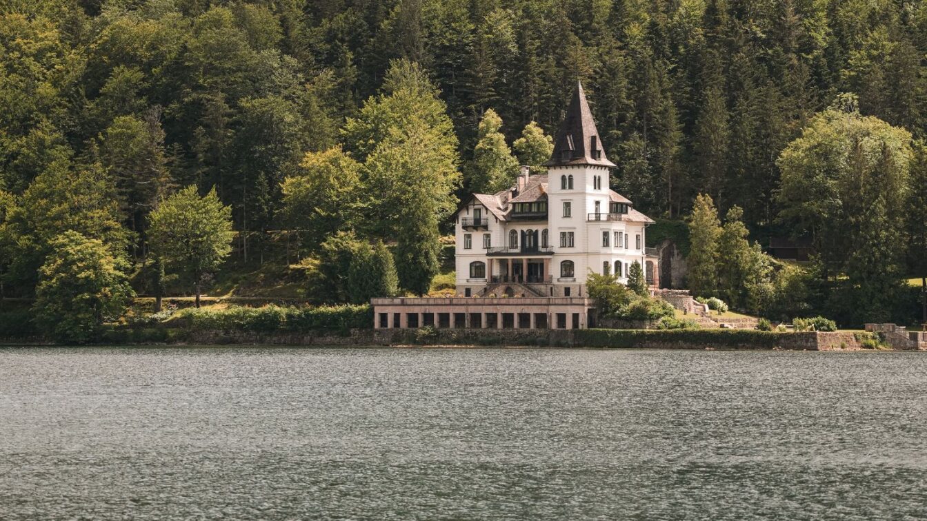 (c) Julia Riedesser | 1000things Historische Villa mit Turm direkt am Ufer des Altausseer Sees, umgeben von dichter Waldlandschaft – malerisches Anwesen im Salzkammergut, eingebettet in idyllischer Naturkulisse.