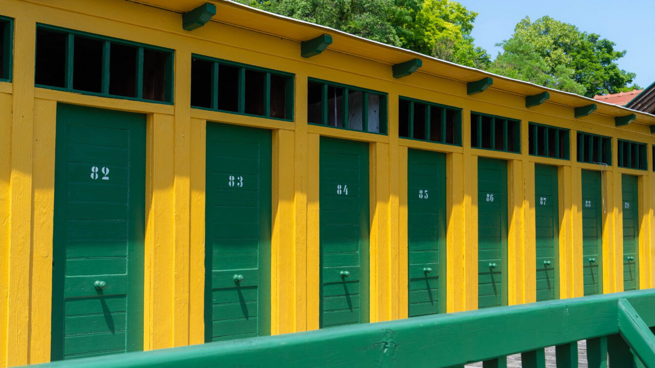 (c) Daniel Leeb | 1000things Creator Community Historische Umkleidekabinen im Thermalbad Bad Fischau – bunte Holzfassade in Gelb und Grün mit nummerierten Türen, nostalgisches Flair im traditionsreichen Freibad in Niederösterreich.