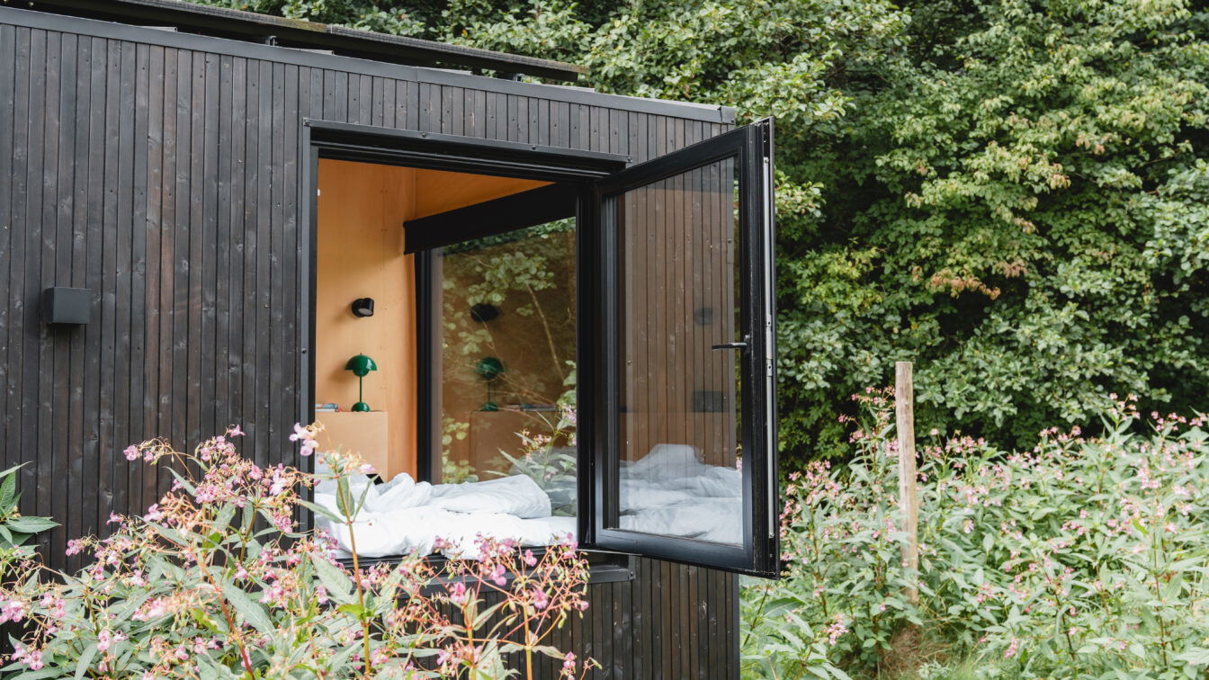 (c) Lilli Wermuth | 1000things Modernes RAUS Tiny House Cabin mit geöffnetem Panoramafenster im Grünen, minimalistisches Design mit Holzverkleidung und Blick ins Schlafzimmer.
