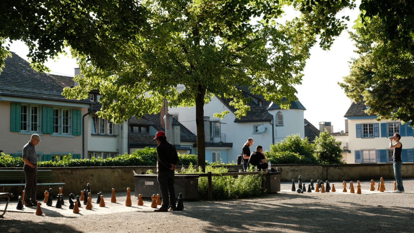 (c) Barbara Palcher | 1000things Menschen spielen im Schatten von Bäumen auf einem großen Outdoor-Schachbrett im Lindenhof in Zürich. Zwei Männer stehen konzentriert am Schachfeld, während andere Personen auf Bänken sitzen und zuschauen. Im Hintergrund sind historische Häuser mit bunten Fensterläden und Schindeldächern zu sehen