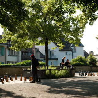 Menschen spielen im Schatten von Bäumen auf einem großen Outdoor-Schachbrett im Lindenhof in Zürich. Zwei Männer stehen konzentriert am Schachfeld, während andere Personen auf Bänken sitzen und zuschauen. Im Hintergrund sind historische Häuser mit bunten Fensterläden und Schindeldächern zu sehen