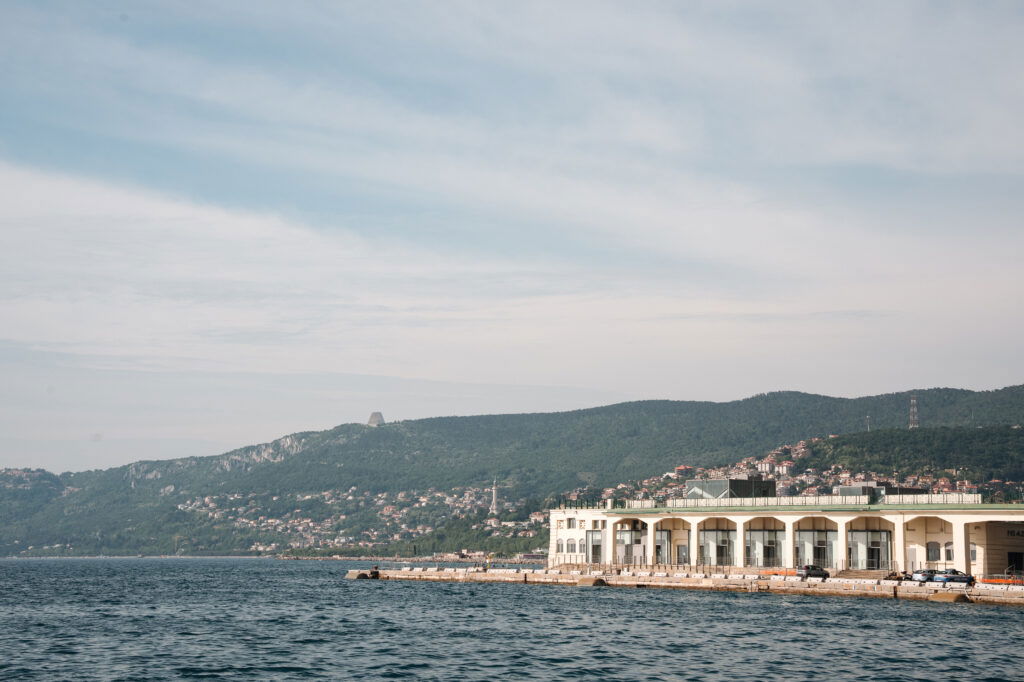 Blick auf die Küste von Triest mit dem historischen Hafenlagerhaus, azurblauem Meer und grünen Hügeln im Hintergrund – mediterrane Hafenstadt mit italienischem Charme und maritimem Flair.