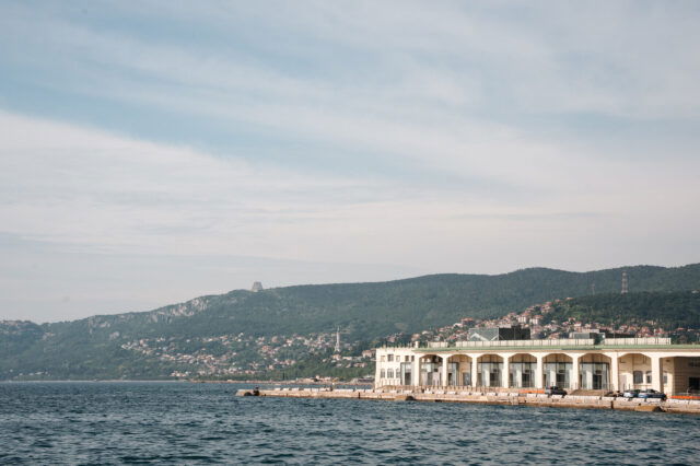 Blick auf die Küste von Triest mit dem historischen Hafenlagerhaus, azurblauem Meer und grünen Hügeln im Hintergrund – mediterrane Hafenstadt mit italienischem Charme und maritimem Flair.