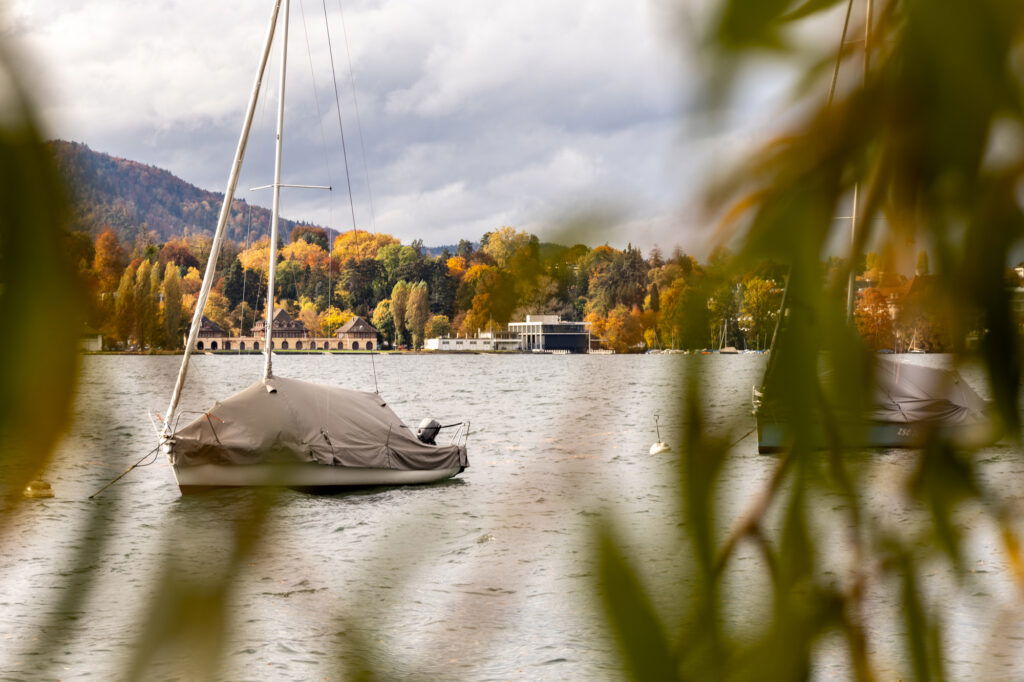 Blick durch herbstlich gefärbte Äste auf ein ruhiges Gewässer mit zwei abgedeckten Segelbooten, die vor Anker liegen. Im Hintergrund sind farbenfrohe Bäume, Gebäude am Ufer und bewaldete Hügel unter einem bewölkten Himmel zu sehen.“