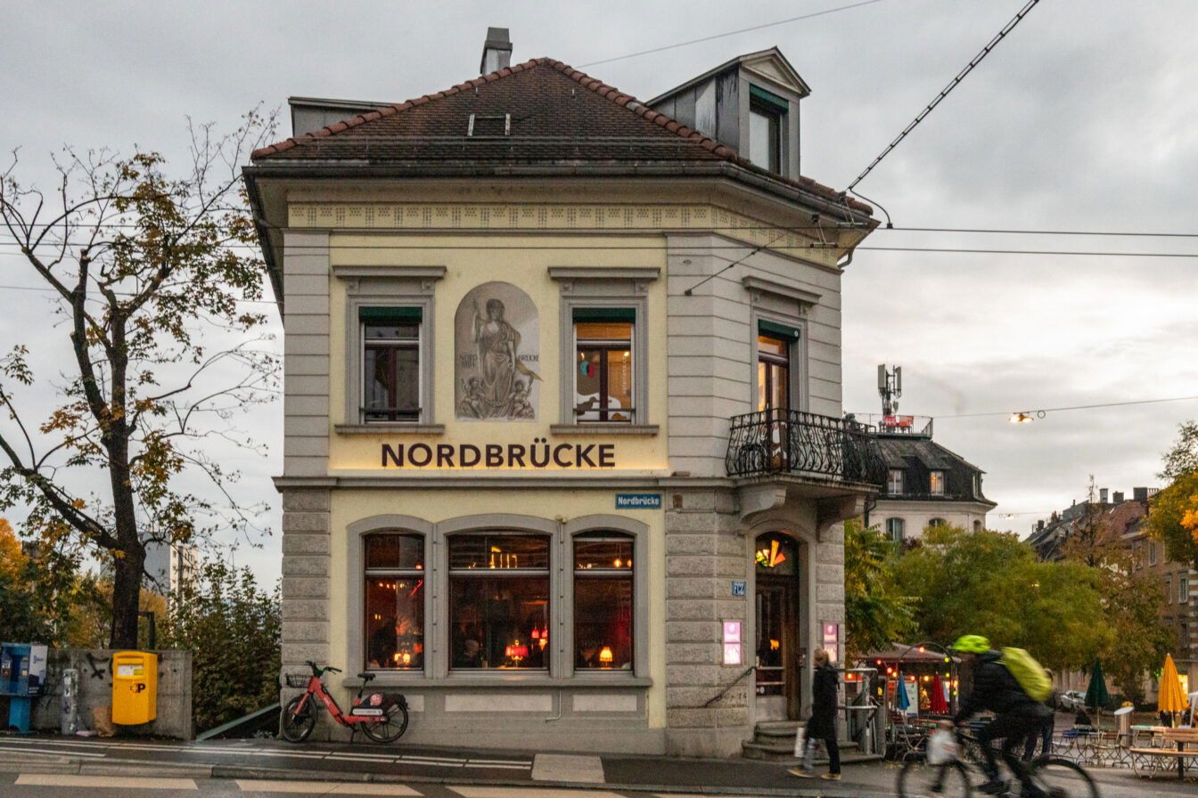 Straßenszene in Zürich mit dem Café-Restaurant Nordbrücke in einem historischen Eckgebäude. Im Vordergrund fährt ein Radfahrer vorbei, während eine Person zu Fuß das Lokal betritt. Die Umgebung ist herbstlich, mit Bäumen und Außentischen auf dem Platz neben dem Gebäude