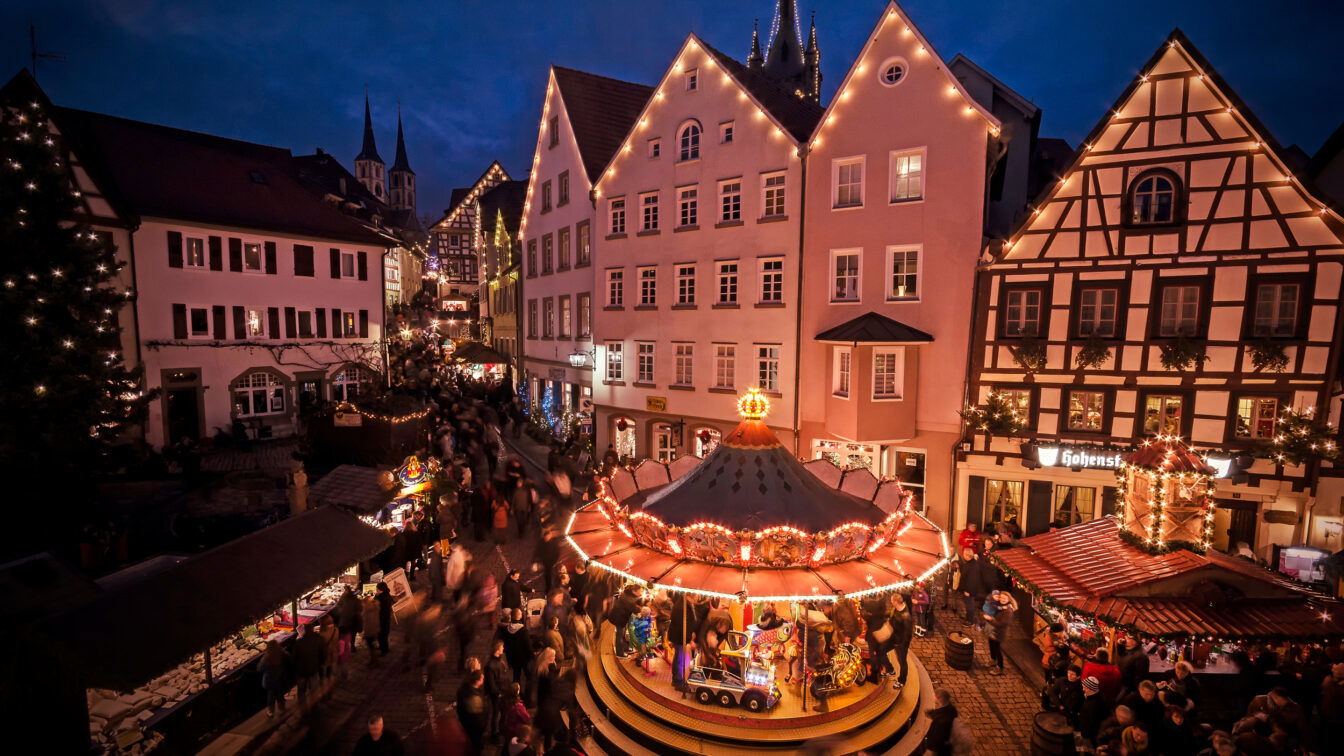 (c) Stadt Bad Wimpfen Das Bild zeigt einen festlich beleuchteten Weihnachtsmarkt in einer historischen Altstadt am Abend. Im Vordergrund befindet sich ein traditionelles Kinderkarussell mit bunten Lichtern und mehreren Figuren, darunter ein Zug und Pferde. Rund um den Platz sind dekorierte Fachwerkhäuser und weitere Stände des Weihnachtsmarkts zu sehen, die von Menschen besucht werden. Im Hintergrund ragen Kirchtürme in den Abendhimmel, während die Atmosphäre von Lichtern und weihnachtlicher Stimmung geprägt ist