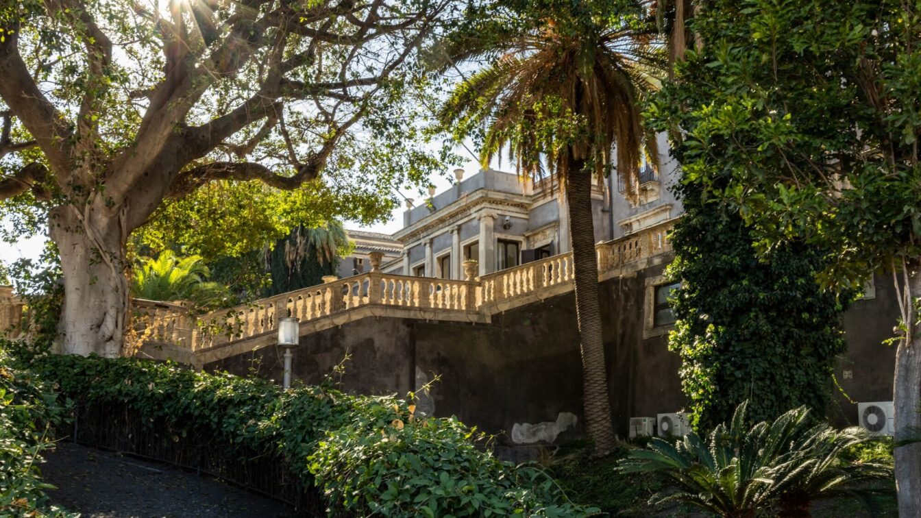 (c) Sonja Koller | 1000things Ein prachtvolles historisches Gebäude in Catania, umgeben von üppigem Grün und Palmen. Sonnenstrahlen brechen durch die Bäume, was der Szene eine friedliche Atmosphäre verleiht.
