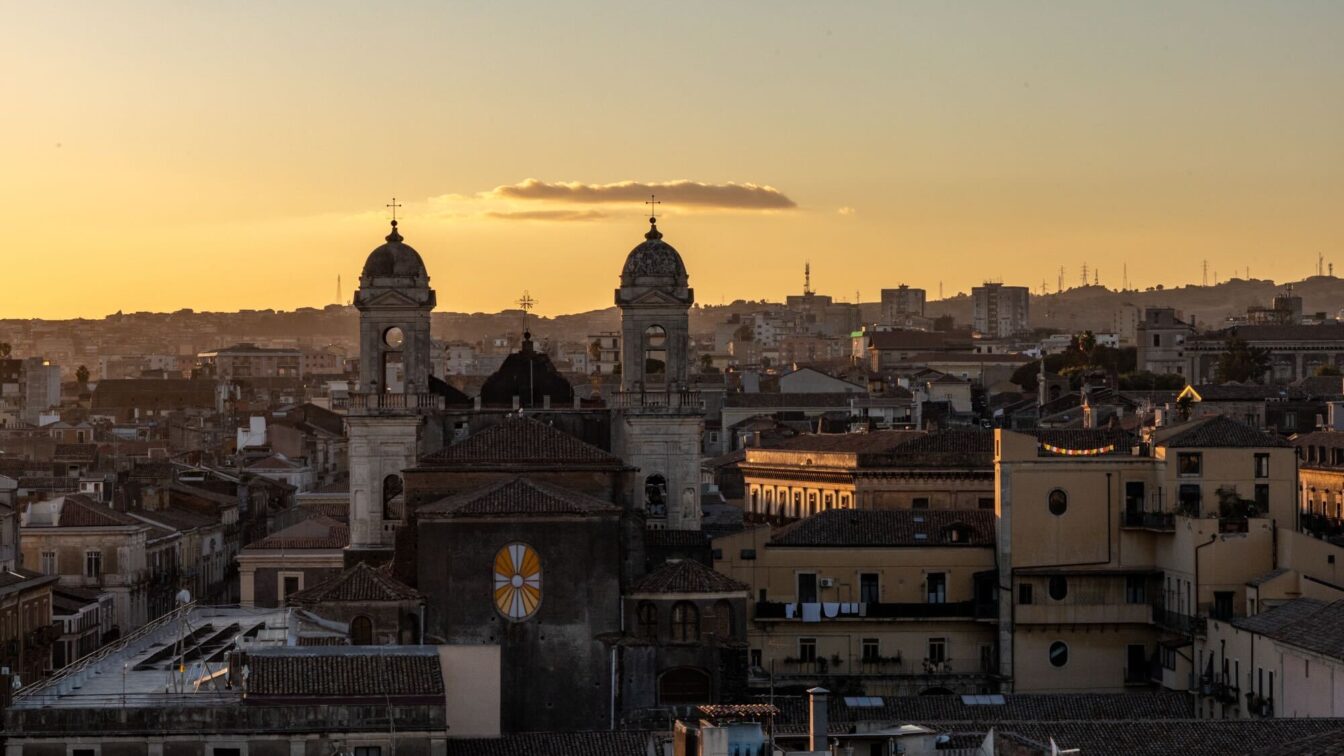 (c) Sonja Koller | 1000things Ein Blick auf Catania, Italien, bei Sonnenuntergang. Die Silhouette zweier Kirchtürme dominiert den Vordergrund, während die Stadt im warmen Abendlicht leuchtet.