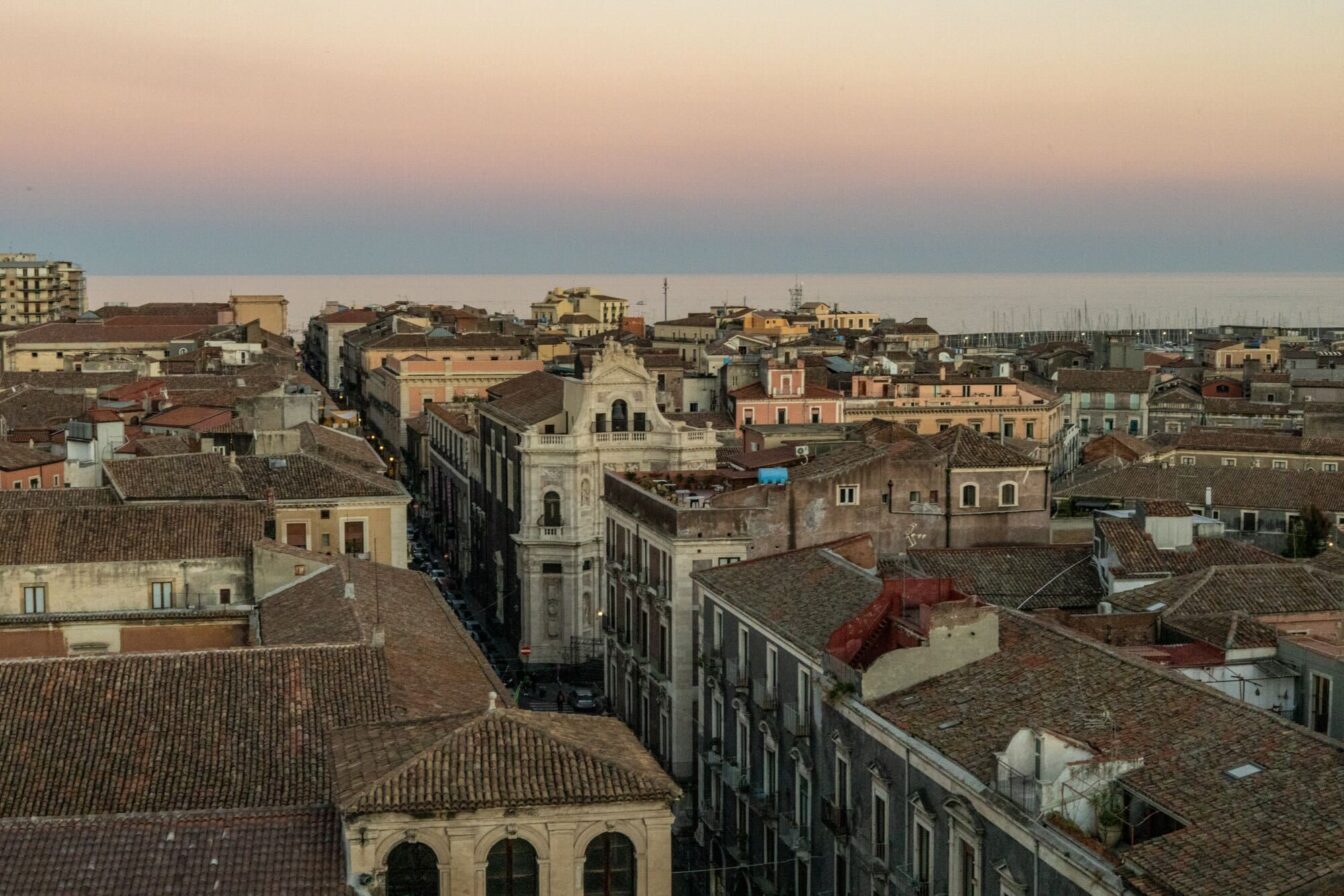Ein Luftbild von Catania, Italien, mit seinen engen, historischen Straßen und ziegelroten Dächern. Der Himmel zeigt zarte rosa und violette Töne in der Abenddämmerung.