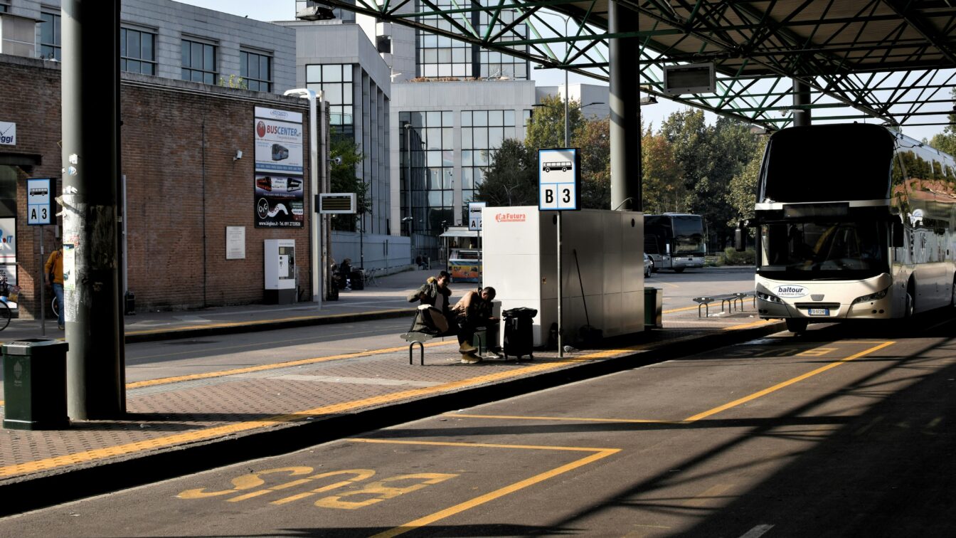 (c) Matteo Maretto | Unsplash Das Bild zeigt eine Busstation an einem sonnigen Tag. Im Vordergrund befindet sich ein leerer Busparkplatz, auf dem der Schriftzug "BUS" gelb auf den Asphalt gemalt ist. Rechts steht ein großer weißer Reisebus mit einem Schriftzug "baltour" an der Front. Daneben sitzen zwei Personen auf einer Bank unter einer Überdachung und warten. Hinter ihnen befinden sich moderne Gebäude mit großen Glasfenstern. Die Szenerie wirkt ruhig, es gibt keine sichtbare Hektik oder Menschenmenge. Schilder mit Busnummern (z.B. B3) kennzeichnen die Abfahrtsbereiche.