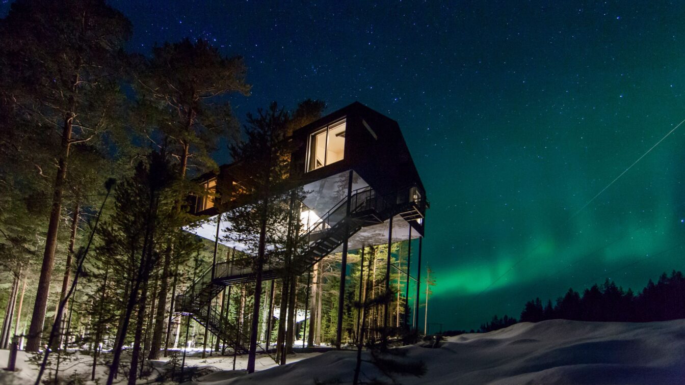 (c) Treehotel Schweden Das Bild zeigt ein modernes Baumhaus-Hotel, das hoch auf Stelzen inmitten eines winterlichen Waldes in Schweden steht. Der Nachthimmel ist mit Sternen bedeckt und die Nordlichter leuchten in intensiven Grüntönen. Eine Außentreppe führt zum erhöhten Eingang des Gebäudes, das teilweise von Bäumen umgeben ist. Die Szenerie vermittelt eine ruhige, abgelegene Atmosphäre in der Natur.