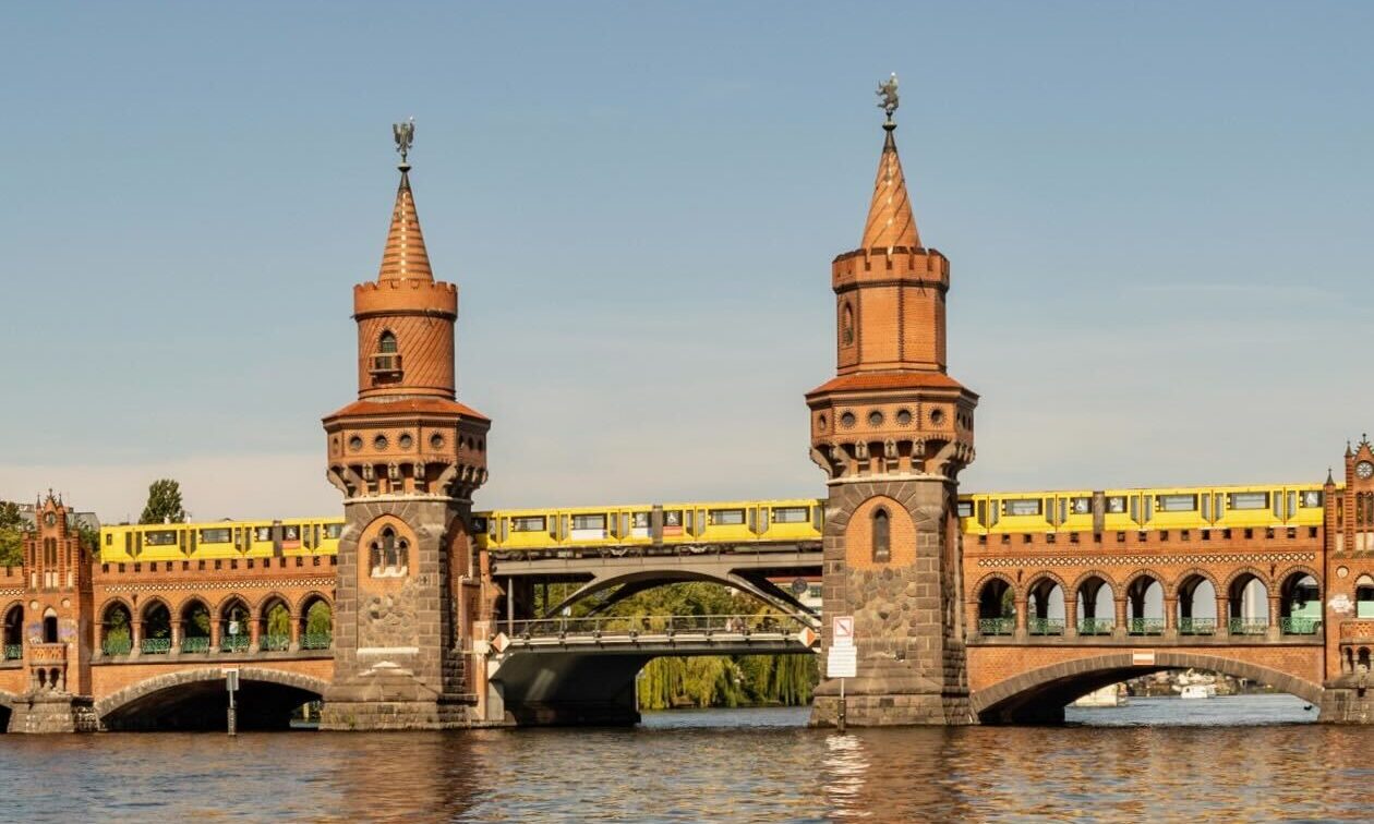 (c) Sonja Koller | 1000things Das Bild zeigt die Oberbaumbrücke in Berlin, eine historische Backsteinbrücke über die Spree mit zwei markanten Türmen. Eine gelbe U-Bahn fährt auf der oberen Ebene der Brücke, während die untere Ebene für Fußgänger und Autos genutzt wird. Im Vordergrund fließt die Spree ruhig unter der Brücke.