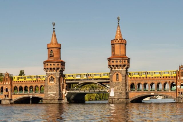 Das Bild zeigt die Oberbaumbrücke in Berlin, eine historische Backsteinbrücke über die Spree mit zwei markanten Türmen. Eine gelbe U-Bahn fährt auf der oberen Ebene der Brücke, während die untere Ebene für Fußgänger und Autos genutzt wird. Im Vordergrund fließt die Spree ruhig unter der Brücke.