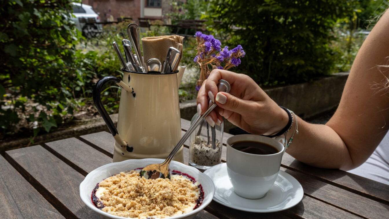 Gemütlicher Brunch im Grünen in Berlin: Eine Hand löffelt ein Dessert mit Beerenkompott und Streuseln, daneben steht eine Tasse Kaffee auf einem Holztisch. Im Hintergrund frisches Besteck in einem nostalgischen Krug und dezente Deko mit Blumen – perfekter Ort für ein entspanntes Frühstück oder Brunch. Ideal zum Reservieren für Wochenenden in Berlin.
