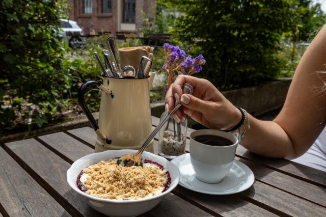 Gemütlicher Brunch im Grünen in Berlin: Eine Hand löffelt ein Dessert mit Beerenkompott und Streuseln, daneben steht eine Tasse Kaffee auf einem Holztisch. Im Hintergrund frisches Besteck in einem nostalgischen Krug und dezente Deko mit Blumen – perfekter Ort für ein entspanntes Frühstück oder Brunch. Ideal zum Reservieren für Wochenenden in Berlin.