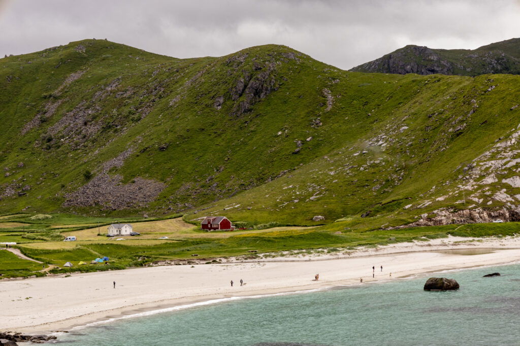 Weitläufige grüne Hügel mit Felsformationen steigen hinter einem hellen Sandstrand an. Einige kleine Häuser, darunter ein rotes Holzhaus und ein weißes Haus, liegen zwischen Wiesen am Fuß der Hügel. Menschen spazieren am Strand entlang, und das türkisfarbene Wasser trifft sanft auf die Küste unter einem bewölkten Himmel.“