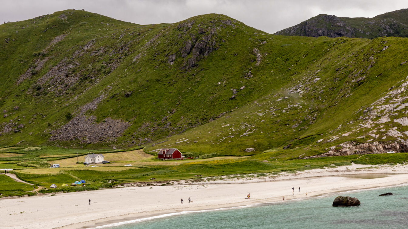 Weitläufige grüne Hügel mit Felsformationen steigen hinter einem hellen Sandstrand an. Einige kleine Häuser, darunter ein rotes Holzhaus und ein weißes Haus, liegen zwischen Wiesen am Fuß der Hügel. Menschen spazieren am Strand entlang, und das türkisfarbene Wasser trifft sanft auf die Küste unter einem bewölkten Himmel.“