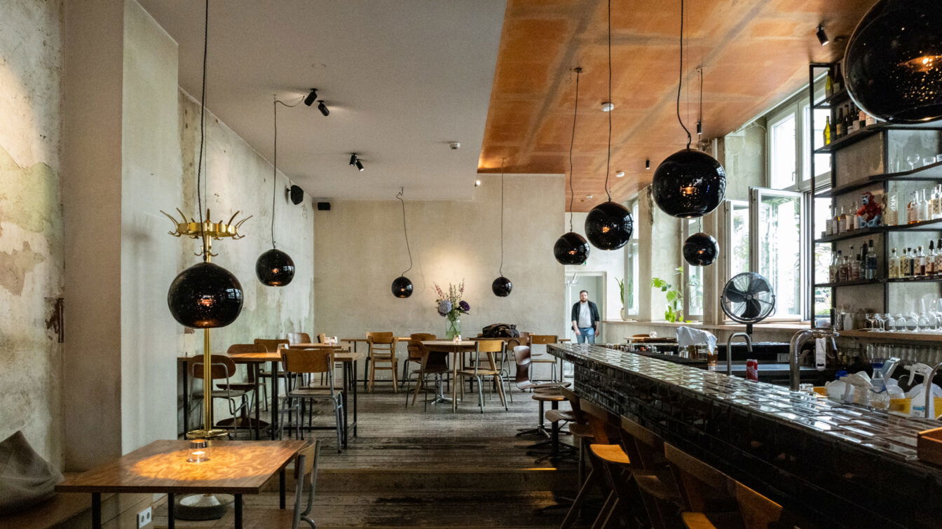 (c) Sonja Koller | 1000things Innenansicht des stilvollen Michelberger Hotels in Berlin: Das Restaurant oder die Bar besticht durch modernes, urbanes Design mit rustikalem Holzboden, schwarzen Hängelampen, offener Bar und kreativem Mix aus Industrial- und Vintage-Elementen. Eine entspannte Atmosphäre für Gäste mit Sinn für Design und Genuss.