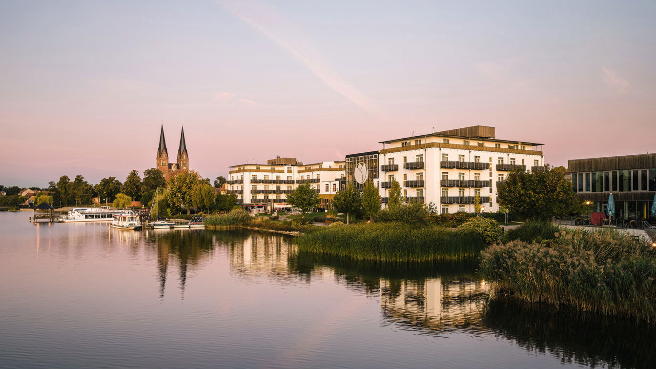 Blick auf das Resort Mark Brandenburg am Ufer eines ruhigen Sees in der Abenddämmerung. Die weißen Gebäude des Resorts reflektieren sich sanft im Wasser, umgeben von üppigem Grün und Schilf entlang der Küste. Im Hintergrund ragen die markanten Kirchtürme eines historischen Gebäudes empor. Der Himmel ist in sanfte Rosa- und Blautöne getaucht, was der Szene eine friedliche, romantische Atmosphäre verleiht.
