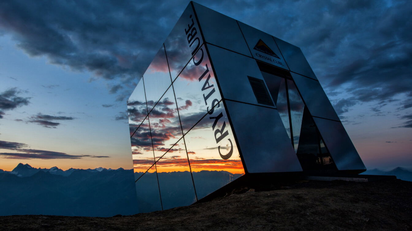 (c) Fisser Bergbahnen GmbH | Andreas Kirschner Das Bild zeigt den Crystal Cube in Serfaus-Fiss-Ladis bei Sonnenuntergang. Der kubusförmige, verspiegelte Bau steht auf einem Berggipfel und reflektiert das atemberaubende Panorama der umliegenden Alpen. Der Himmel ist von dramatischen Wolken bedeckt, die sich in den Spiegelwänden des Crystal Cube widerspiegeln. Die warmen Farben des Sonnenuntergangs kontrastieren mit den kühlen Tönen des Himmels und verleihen der Szene eine mystische Atmosphäre.