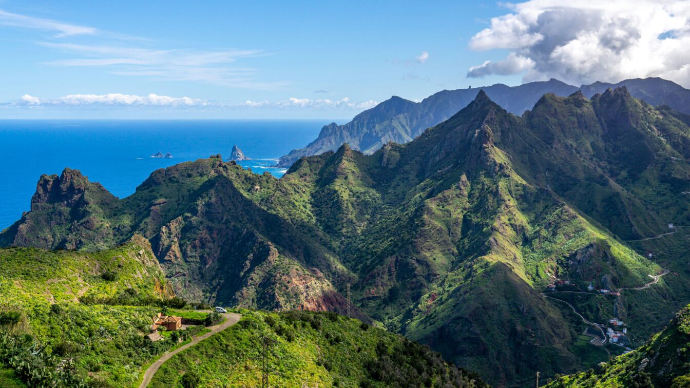 (c) Hendrik Cornelissen | Unsplash Panorama der grünen, zerklüfteten Berglandschaft auf Teneriffa, Kanarische Inseln. Im Vordergrund eine kleine Straße und ein Haus, während die bewachsenen Berge sich zum Meer hin erstrecken. Am Horizont liegt der blaue Atlantik, gesäumt von Felsen im Wasser und sanften Wolken am Himmel.
