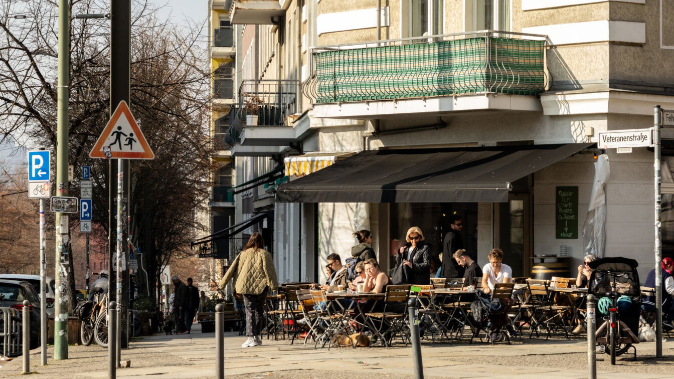 Ein typischer Berliner Straßenzug mit einer einladenden Café- und Bar-Szene. Die Szene spielt sich an der Veteranenstraße ab, wo Menschen das sonnige Wetter genießen und sich an den Tischen eines Straßencafés niederlassen. Schwarze Markisen spenden Schatten, während die Gäste in entspannter Atmosphäre sitzen, plaudern und ihr Essen oder Getränke genießen.