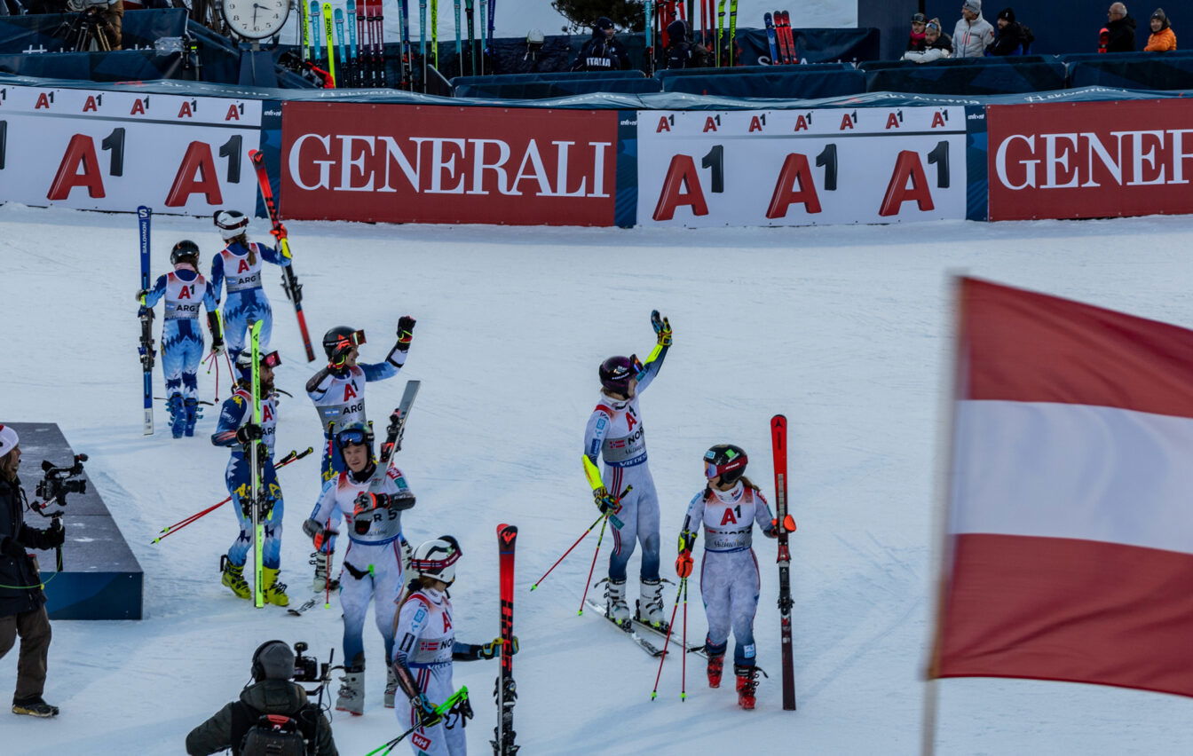 Mehrere Skirennläufer*innen in Rennanzügen stehen nach einem alpinen Skirennen im Zielbereich auf der Piste, einige winken ins Publikum; im Hintergrund sind Werbebanden zu sehen, im Vordergrund weht eine österreichische Flagge.
