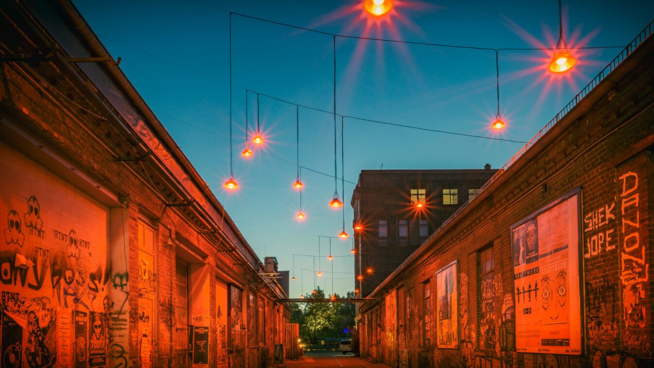 (c) Stephan Widua | Unsplash Eine schmale Gasse zwischen alten Backsteingebäuden mit Graffiti, beleuchtet von orangefarbenen Lampen, die stimmungsvoll im Abendlicht hängen.