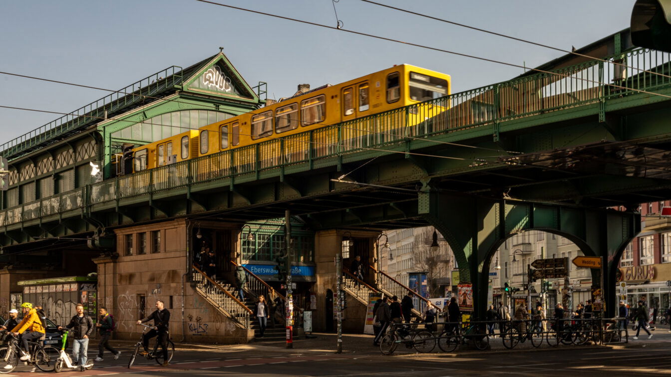 (c) Sonja Koller | 1000things Die U-Bahn-Station Eberswalder Straße in Berlin mit einer gelben U-Bahn, die über eine grüne Stahlbrücke fährt. Die historische Hochbahnstation hat eine markante Architektur mit verzierten Fenstern und einem Giebeldach mit Graffiti. Unter der Brücke beleben Fußgänger, Radfahrer und Autos die Kreuzung. Mehrere Menschen steigen die Treppen zur Station hinauf oder verlassen sie. Im Hintergrund sind Geschäfte, Cafés und Werbetafeln zu sehen, während die tief stehende Sonne lange Schatten auf die Szene wirft