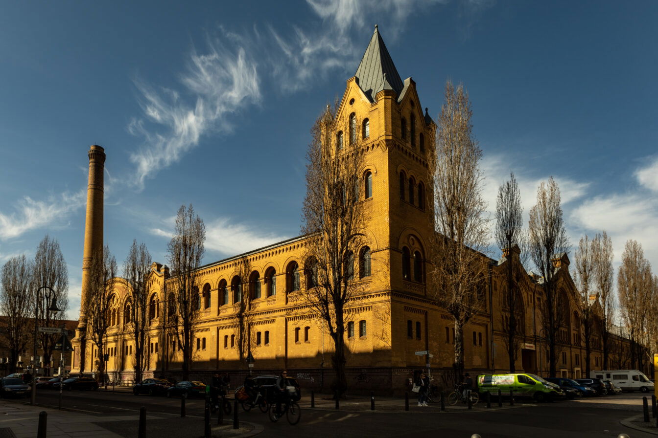 Die Kulturbrauerei in Berlin-Prenzlauer Berg, ein beeindruckendes historisches Backsteingebäude mit neoromanischer Architektur. Die gelblich-braunen Ziegelwände sind mit Rundbogenfenstern und verzierten Elementen gestaltet. Ein hoher Schornstein ragt links in den Himmel, während rechts ein Turm mit spitzem Dach das Gebäude dominiert. Vor dem Gebäude stehen mehrere hohe, kahle Bäume, deren Schatten auf die Fassade fallen. Auf der Straße davor sind Autos, Fahrräder und Fußgänger zu sehen, die sich durch das belebte Stadtviertel bewegen. Der Himmel ist blau mit dekorativen, wellenförmigen Wolken, die eine malerische Kulisse für die Szene bilden.
