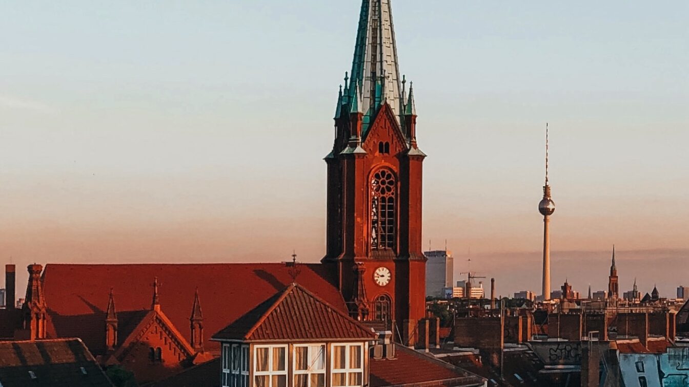 (c) Sonja Koller | 1000things Blick auf die Gethsemanekirche im Berliner Stadtteil Prenzlauer Berg bei Sonnenuntergang mit dem Berliner Fernsehturm im Hintergrund – architektonisches Highlight im Prenzlauer Berg Guide und beliebter Fotospot in Berlin.