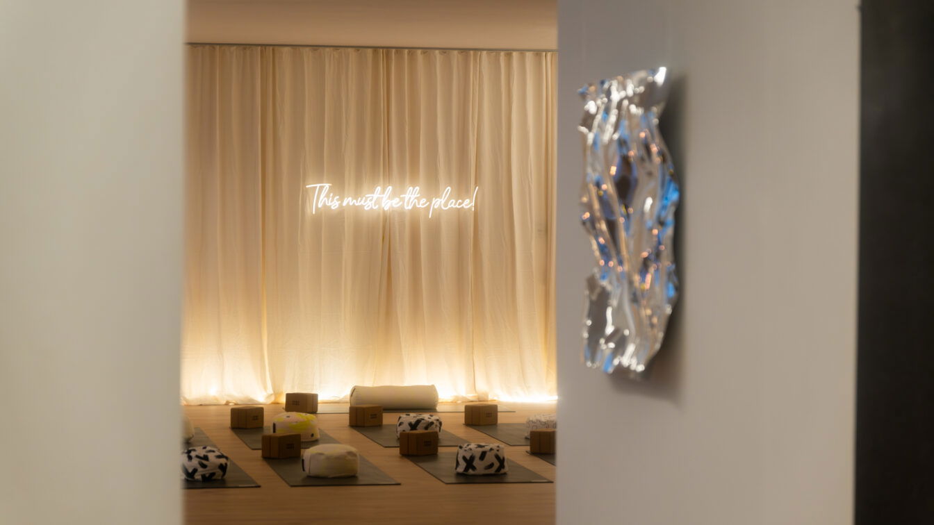 SHA-LA Studio Prenzlauer Berg | (c) Julia Rosenau Eingang zu einem Yogastudio mit auffälliger Lichtstimmung. Am Boden liegen Matten mit Kissen. An der Wand hängt ein Leuchtschild mit der Aufschrift "This must be the place".