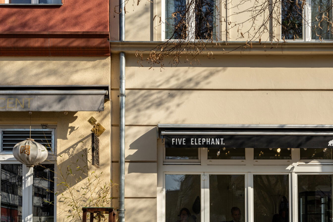 Die Fassade des beliebten Berliner Cafés Five Elephant im Stadtteil Kreuzberg. Die Außenansicht zeigt die charakteristische schwarze Markise mit weißer Schrift, die den Namen des Cafés trägt. Durch die großen Fenster lässt sich ein Blick in den Innenraum erhaschen, wo Gäste bei Kaffee und Gebäck entspannen.

Die Szene ist in warmes Sonnenlicht getaucht, wodurch sich sanfte Schatten der umliegenden Bäume auf der beigen Hauswand abzeichnen. Daneben ist ein Teil eines benachbarten Geschäfts zu sehen, das mit einem asiatisch inspirierten Lampion dekoriert ist. Die ruhige, einladende Atmosphäre spiegelt den Charme dieses Viertels wider, das für seine erstklassige Kaffeehauskultur bekannt ist. Five Elephant ist besonders berühmt für seinen exzellenten Spezialitätenkaffee und den hausgemachten New York Cheesecake, die zu den besten der Stadt zählen.