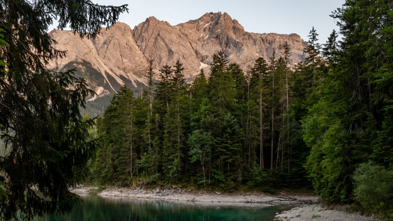 (c) Sonja Koller | 1000things Blick auf die Zugspitze bei Sonnenuntergang vom Eibsee bei Garmisch-Partenkirchen in Bayern – klare Berglandschaft mit grünem Wald und türkisfarbenem Wasser im Vordergrund.